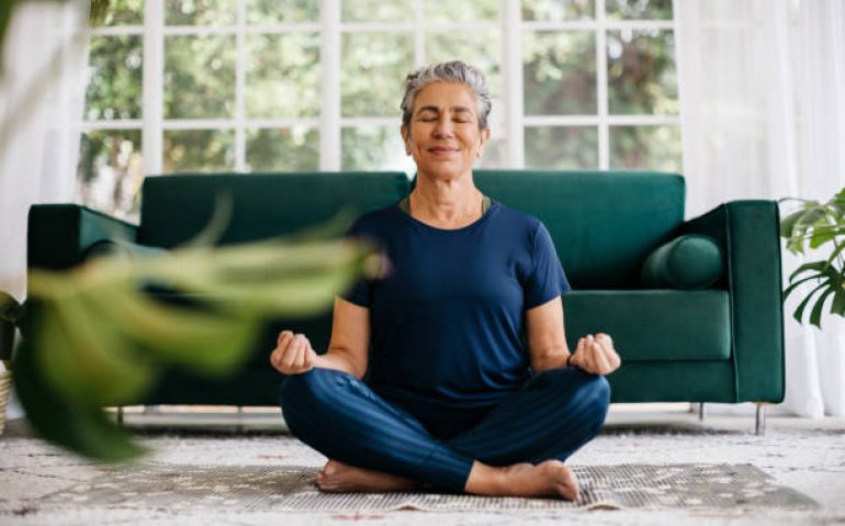 Senior woman meditating in lotus position at home, sitting on the floor in fitness clothing. Mature woman doing a breathing workout to achieve relaxation, peace and mindfulness.